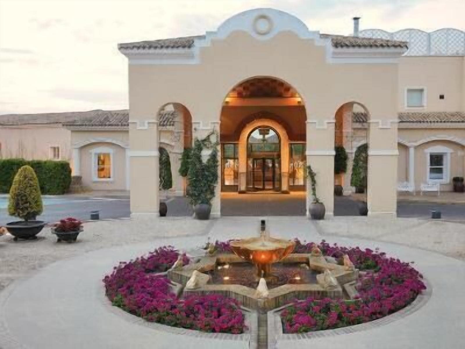 A hotel entrance with an arched doorway, a central fountain, and a landscaped garden with purple flowers.