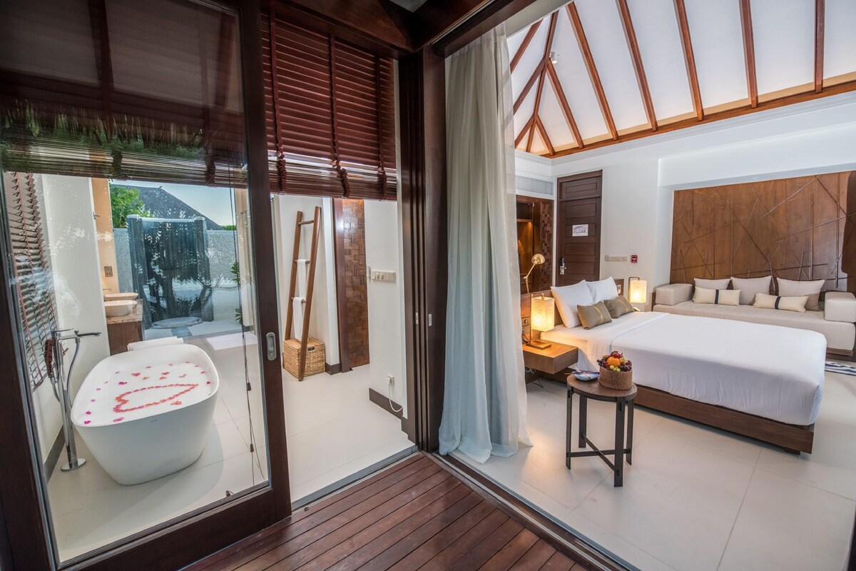 Une chambre moderne avec un grand lit, une table de chevet et une vue sur la salle de bain, accessible par une porte vitrée, où l’on aperçoit une baignoire.
