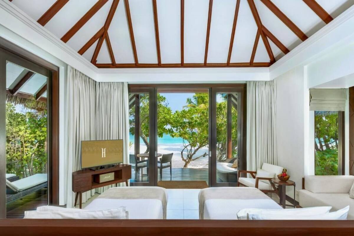 Quarto de hotel com uma grande porta de correr de vidro que dá para a praia, uma cama, uma televisão e um espaço de estar com vista.