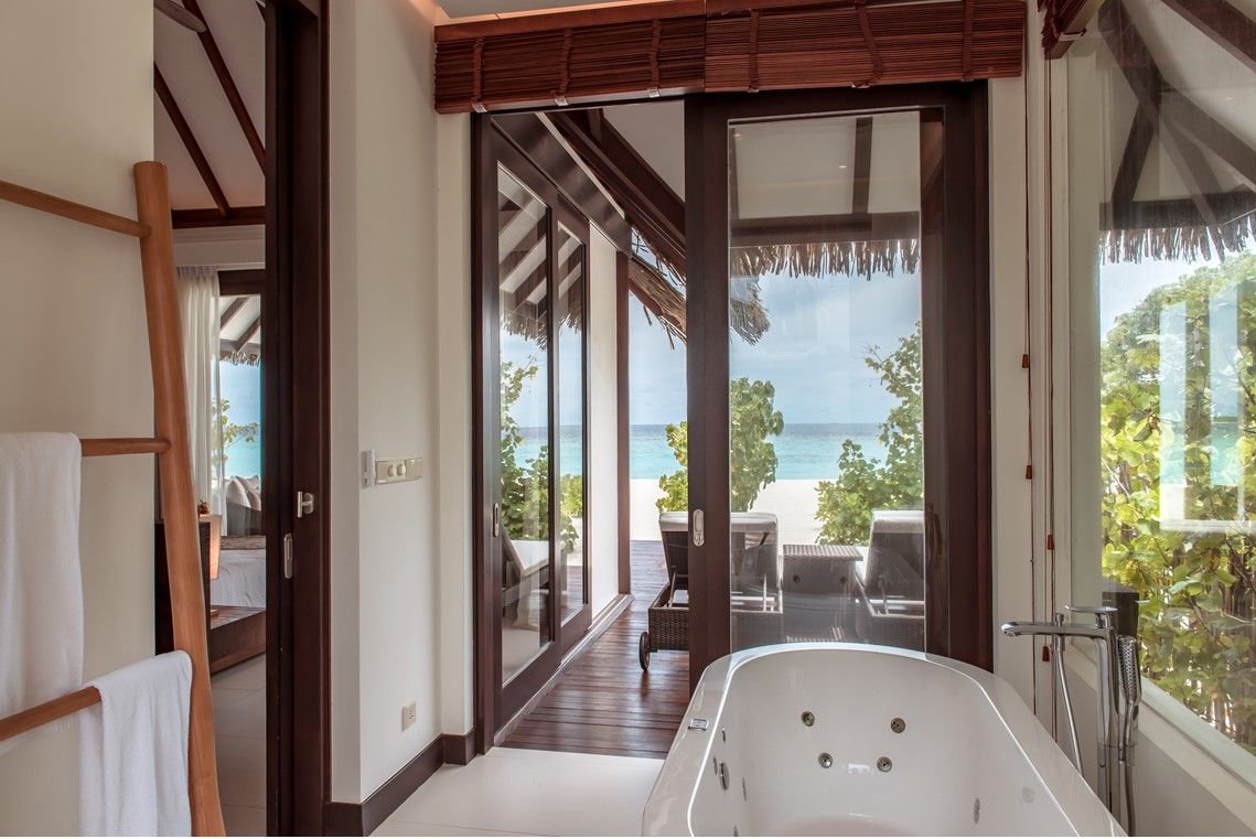 A modern bathroom with a large bathtub, a glass door leading to a balcony with a view of the beach.