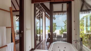 A modern bathroom with a large bathtub, a glass door leading to a balcony with a view of the beach.
