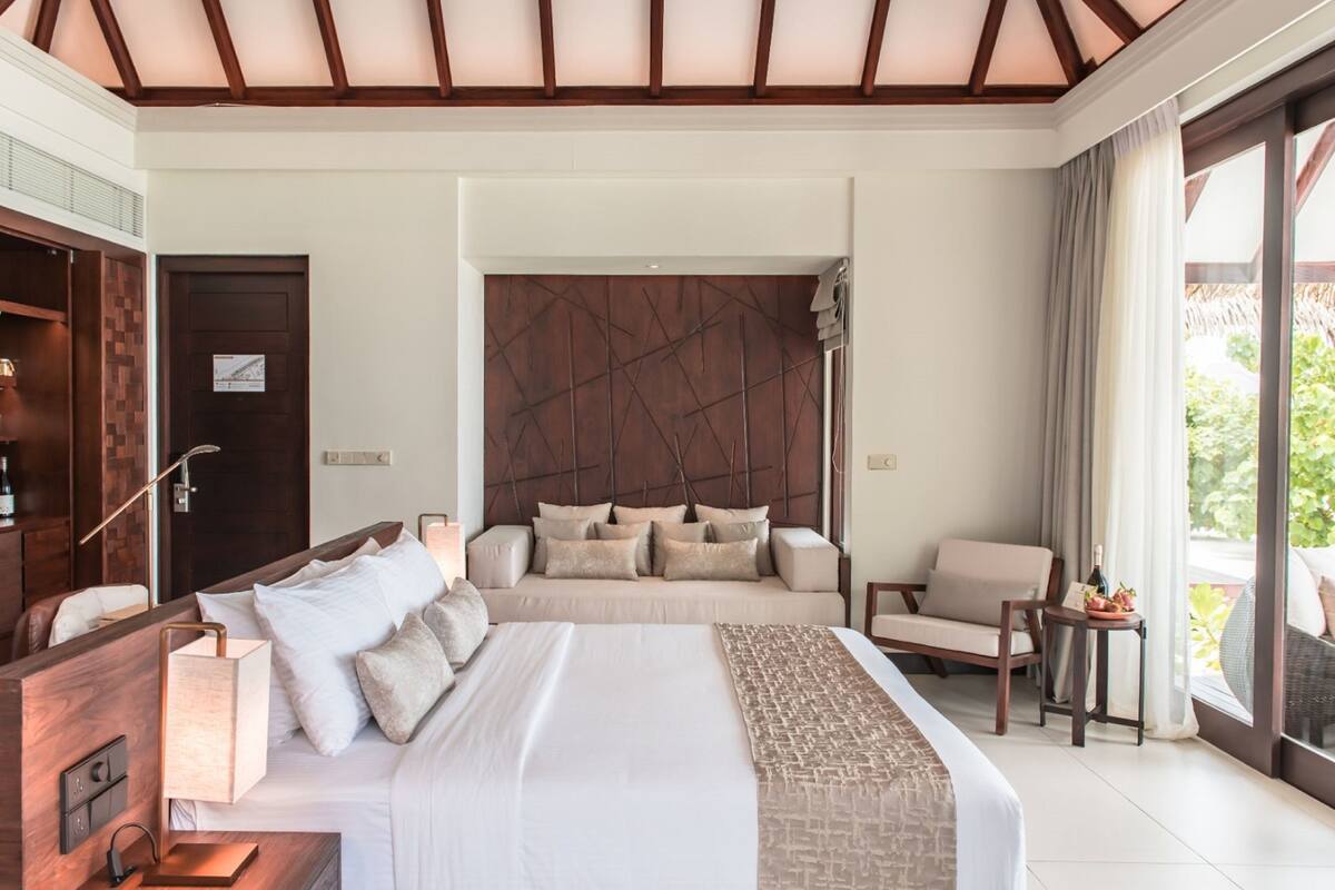Quarto de hotel com uma cama grande, cabeceira de madeira, sofá, poltrona e uma mesinha com uma garrafa e uma planta.