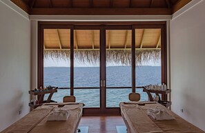 Una habitación con dos camas, un ventanal con vista al mar, techo de madera y una pequeña mesa con artículos de aseo.
