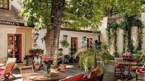 An outdoor patio area with round tables and wooden chairs, surrounded by potted plants and a large tree. - Alchymist Grand Hotel and Spa (Prague)