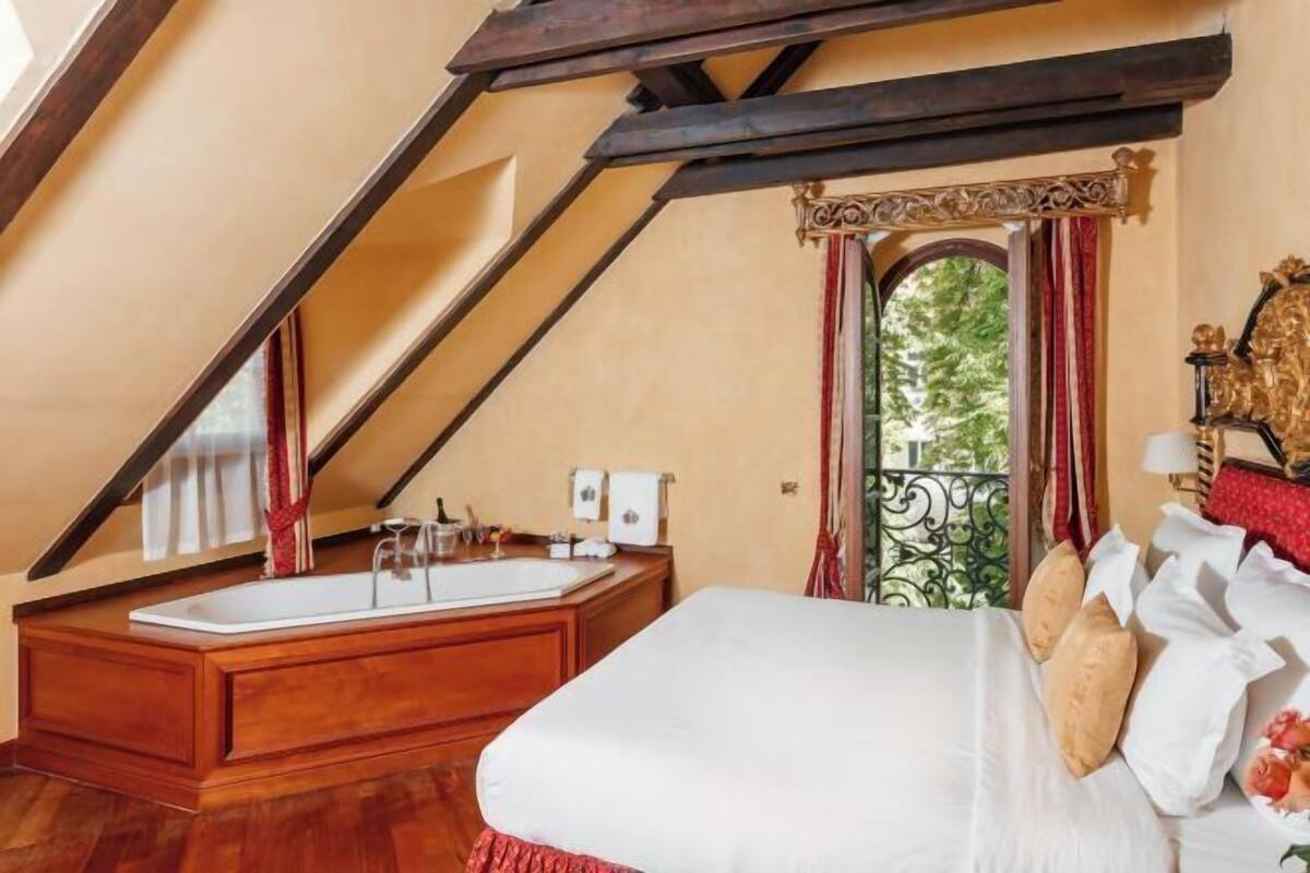 Une chambre à coucher avec un grand lit, une coiffeuse en bois et une baignoire, ainsi qu’un balcon doté d’une balustrade en fer forgé.