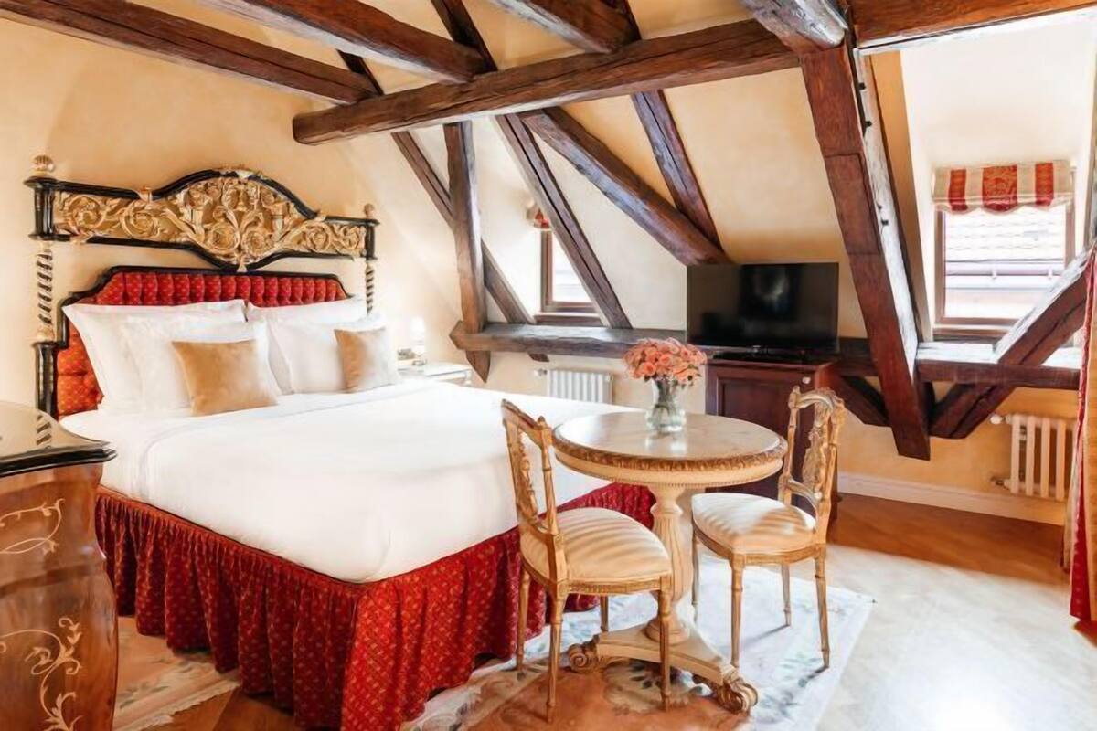 Une chambre à coucher avec un grand lit, une table ronde, deux chaises, un téléviseur et une fenêtre avec des rideaux.