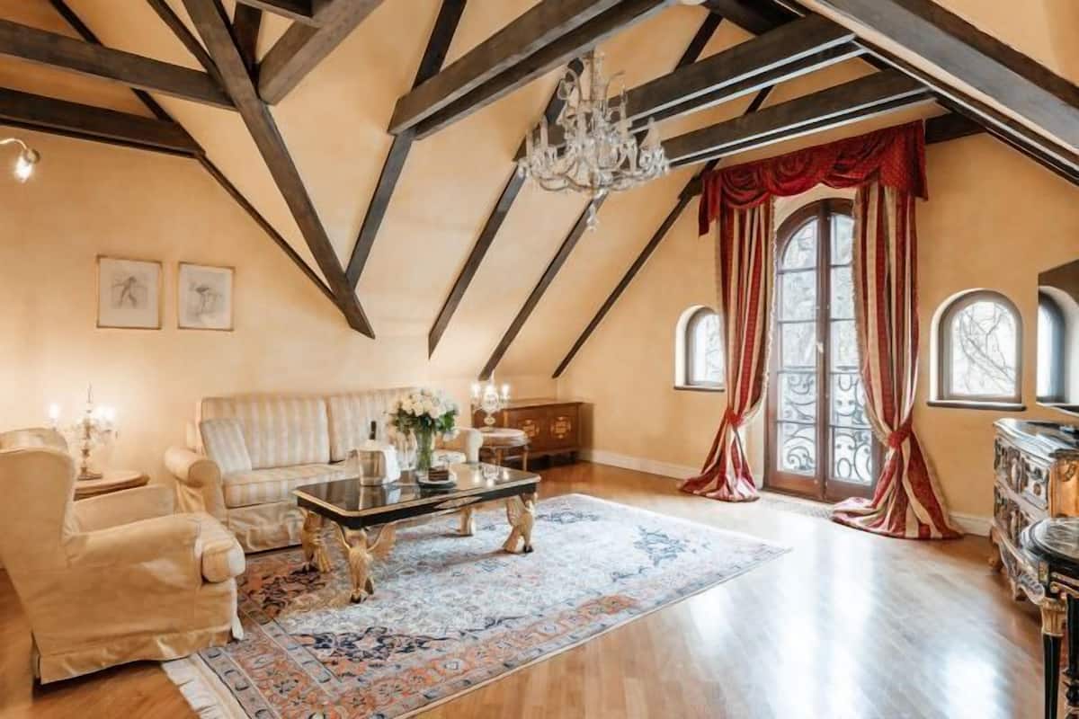 Un salon spacieux avec un plafond haut, des poutres en bois, un lustre, un canapé, une table basse et une grande fenêtre avec des rideaux.