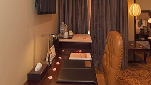 A hotel room with a wooden dining table, a leather chair, a flat-screen TV mounted on the wall, a small table with a bottle and a cup, and a large window with curtains. - Inn And Go Kuwait Plaza Hotel (Kuwait City)
