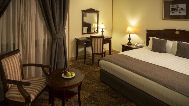A hotel room with a bed, a desk, a chair, a small table with fruit, and a lamp. - Inn And Go Kuwait Plaza Hotel (Kuwait City)