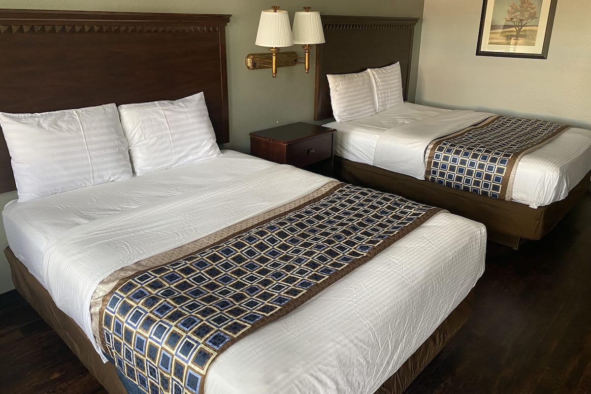 Two double beds with white linens and patterned bedspreads, a nightstand with a lamp, and a framed picture on the wall.