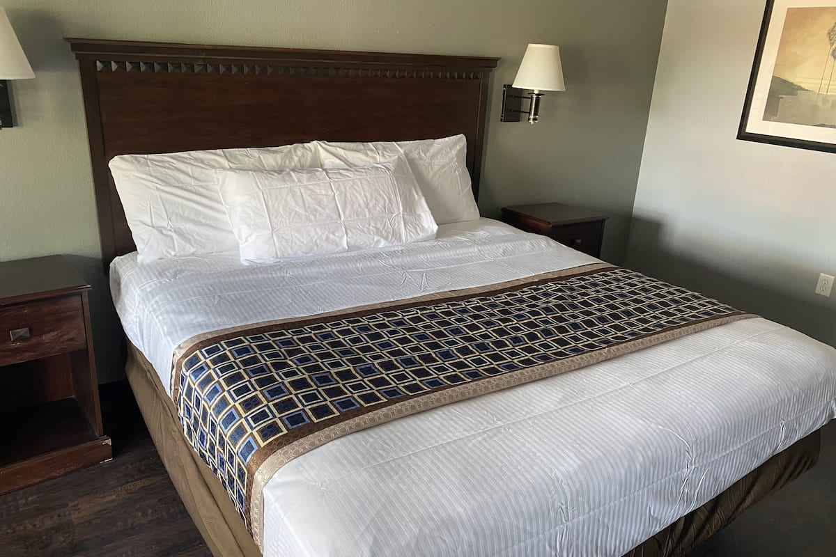 A neatly made bed with white linens and a patterned throw blanket, a wooden headboard, a bedside lamp, and a framed picture on the wall.