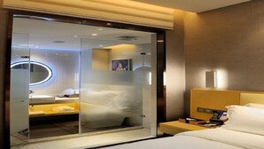 A modern hotel room with a glass-enclosed shower area, a wooden headboard, and a bedside table with a phone and a lamp.