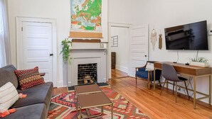 Una sala de estar con chimenea, un sofá, una mesa de centro, un escritorio con silla, y un televisor montado en la pared.
