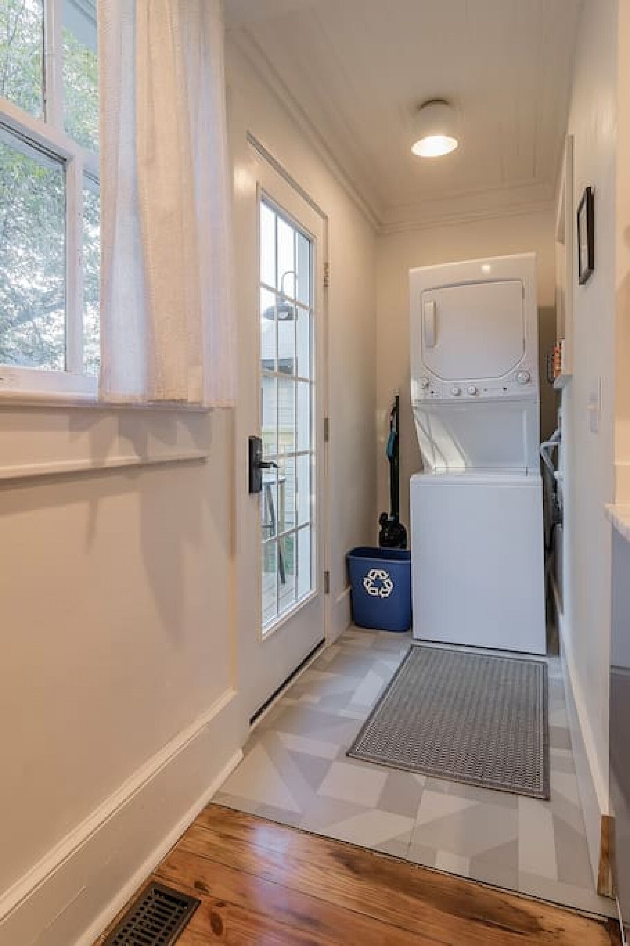 A narrow hallway with a wooden floor, a white door with glass panels, a white washing machine, and a blue recycling bin.