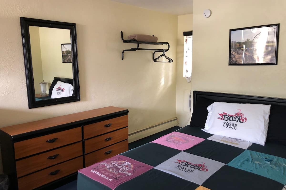A hotel room with a bed, a wooden dresser, a mirror, a wall-mounted shelf, and a framed picture.