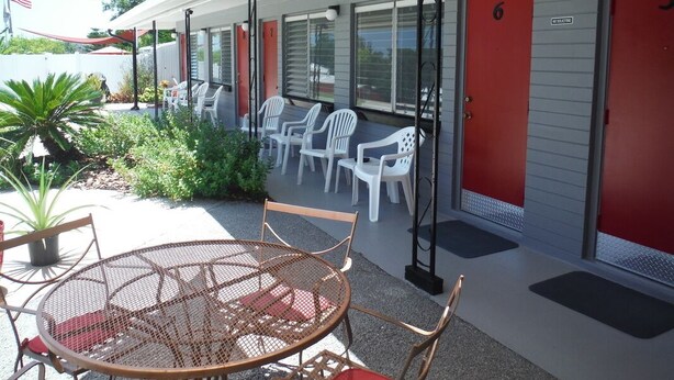 A motel with a covered patio area featuring a round table and chairs, a small garden, and red doors numbered 6 and 7. - The Scoot Inn (Port Orange)