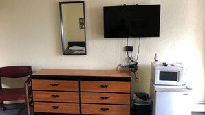 A hotel room with a wooden dresser, a mirror, a television, a microwave, and a trash can. - The Scoot Inn (Port Orange)