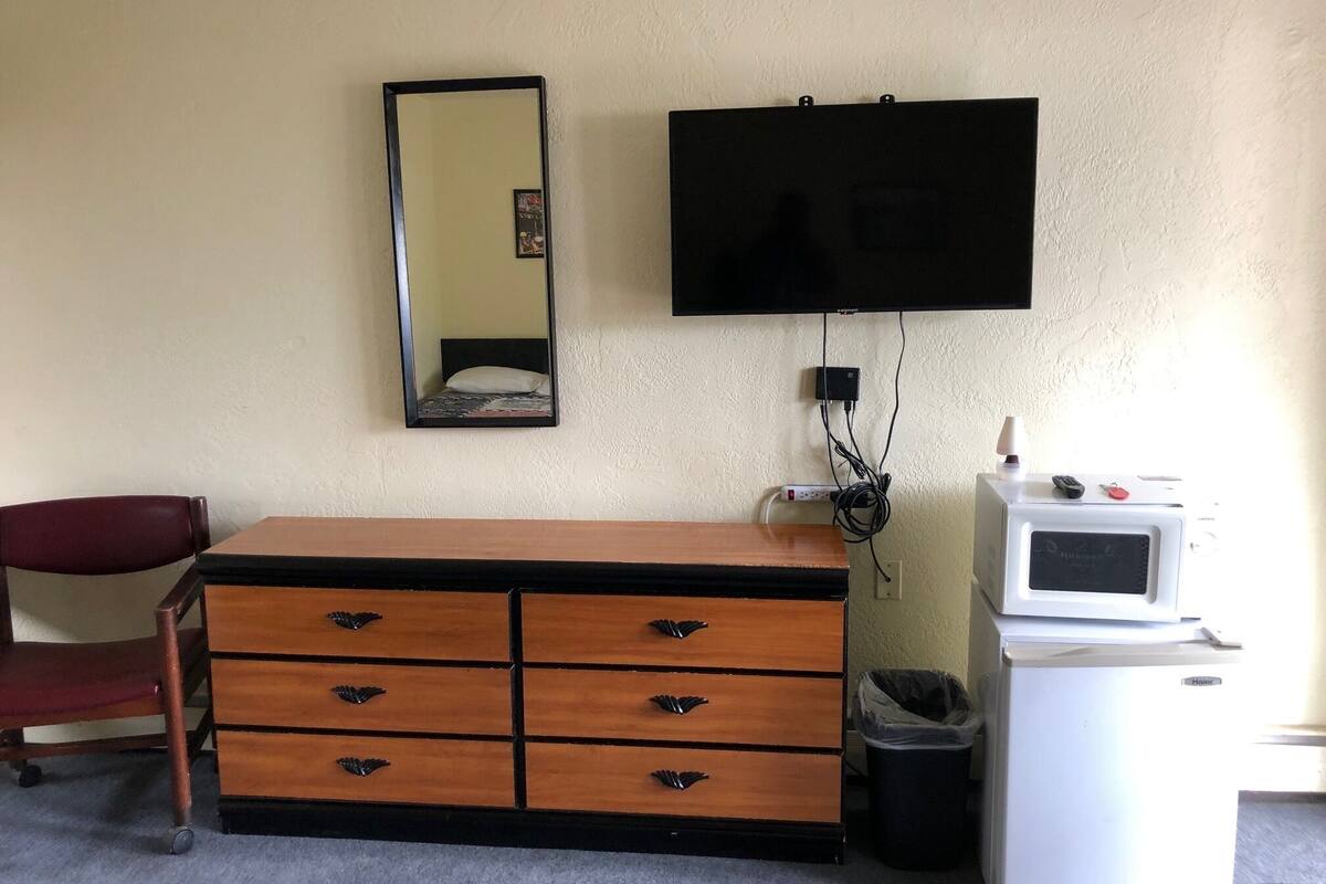A hotel room with a wooden dresser, a mirror, a television, a microwave, and a trash can.