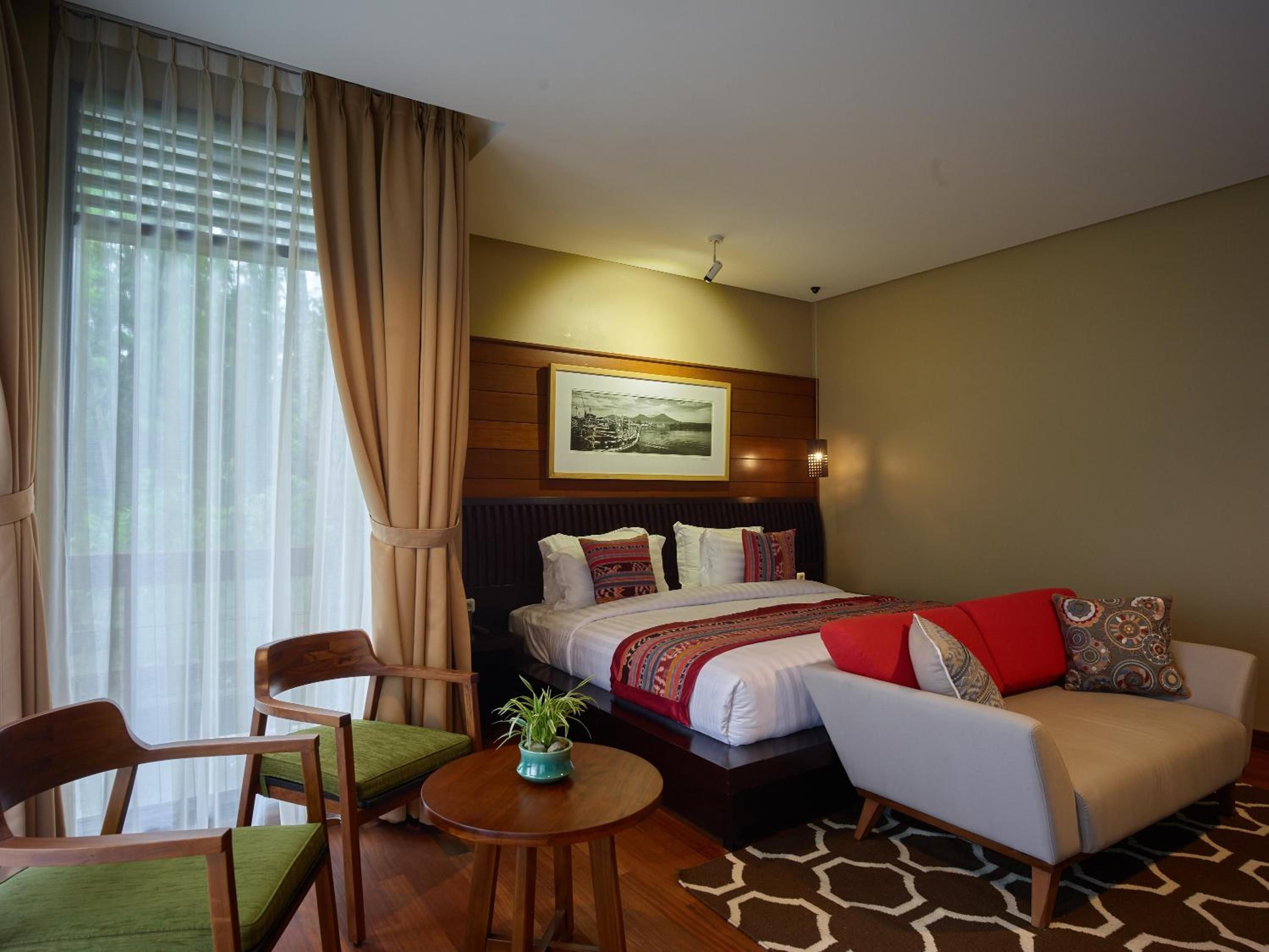 A hotel room with a bed, a sofa, a chair, a small table with a plant, and a large window with curtains.