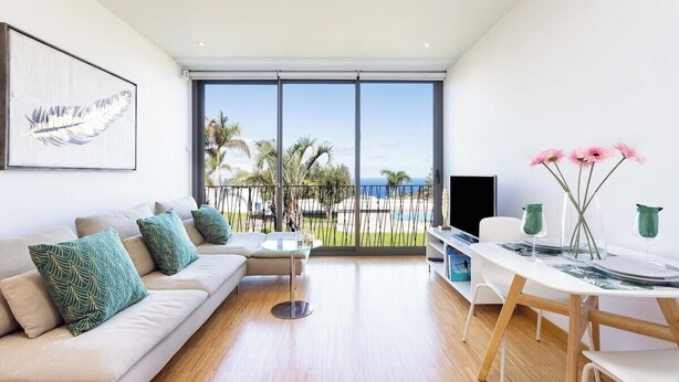 A modern living room with a sofa, coffee table, and dining table. There is a large window with a view of palm trees and a fence.