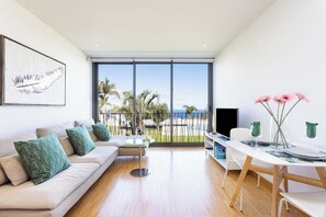 Ein modernes Wohnzimmer mit Sofa, Couchtisch und Esstisch. Ein großes Fenster bietet Blick auf Palmen und einen Zaun.