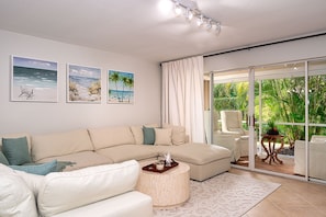 Uma sala de estar moderna com um sofá seccional bege, uma mesa de centro redonda e uma porta de vidro que dá para uma varanda com vista para o verde.