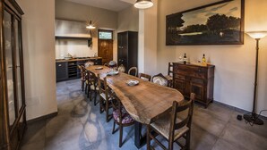 A dining area with a long wooden table, chairs, a painting on the wall, and a kitchen in the background. - Villa del Poeta - Homelike Villas (Loro Ciuffenna)