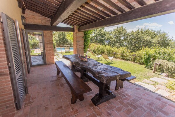 An outdoor patio with a wooden table and benches, a brick floor, and a view of a swimming pool and greenery.