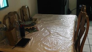 A small dining area with a table covered by a plastic sheet, wooden chairs, and a refrigerator in the background. - Come, Relax and feel at Home (Tijuana)