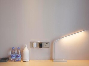 Una camera d'albergo moderna e pulita con un comodino, due bottiglie d'acqua, una bottiglia d'acqua bianca, una presa elettrica e una lampada a parete.