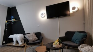 Uma sala de estar moderna com uma TV de tela plana na parede, um sofá, uma poltrona e uma pequena mesa redonda com uma tigela de frutas.