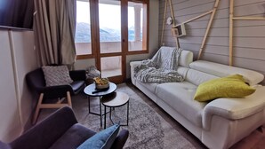 Uma sala de estar moderna com um sofá branco, uma poltrona preta e uma mesa redonda pequena com uma tigela de frutas. Grandes janelas oferecem vista para as montanhas.