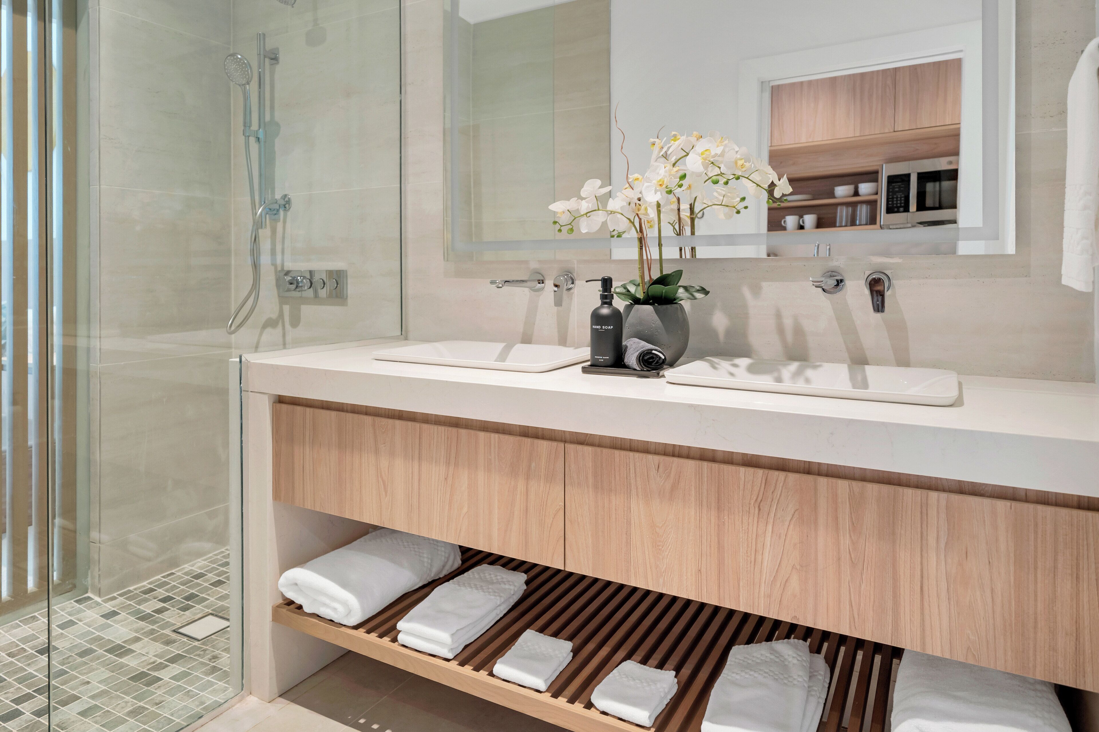 Une salle de bain moderne avec une douche à parois de verre, un meuble-lavabo à deux vasques et une étagère en bois où sont posées des serviettes.