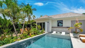 Una piscina con agua cristalina, rodeada de plantas tropicales y una casa con techo de tejas.