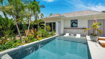 Une piscine à l’eau claire, entourée de plantes tropicales et une maison avec un toit en tuiles.