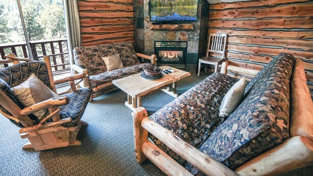 Interior de una cabaña de troncos con chimenea, televisor montado en la pared y un rincón de estar acogedor con un sofá y sillones estampados.