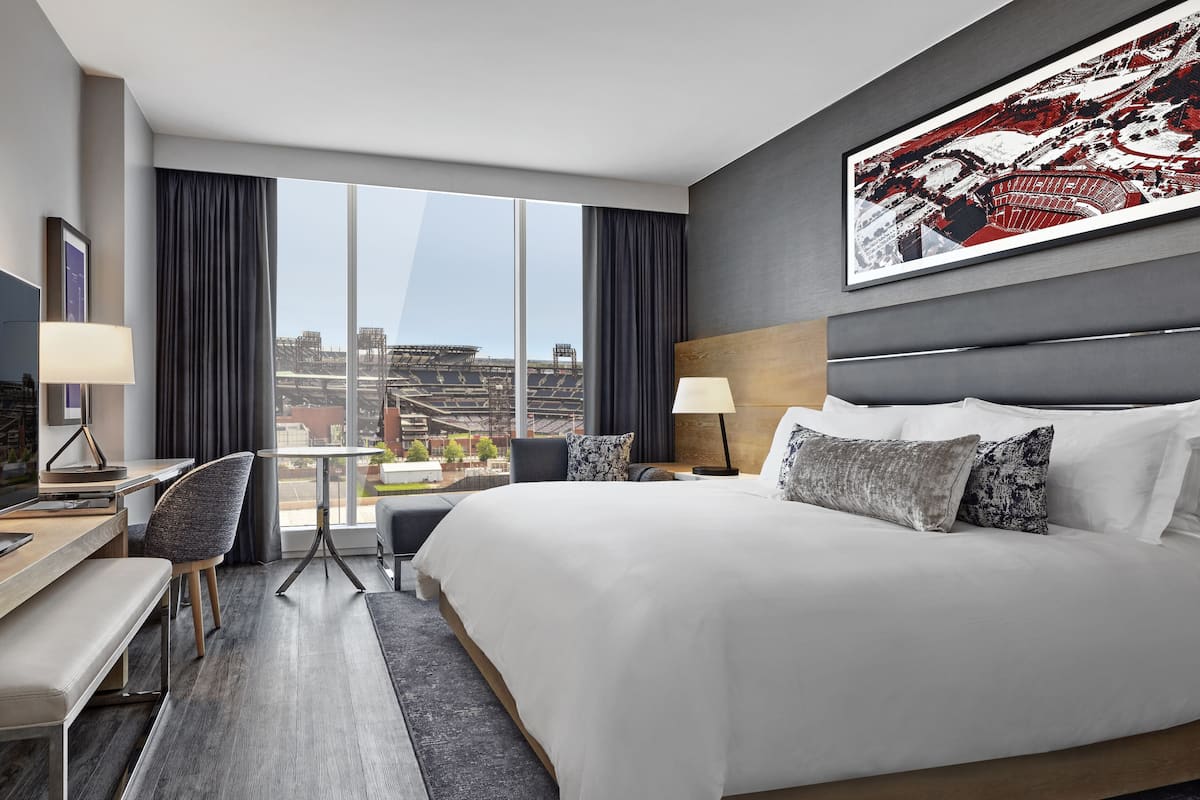 A modern hotel room with a large bed, a desk with a chair, a TV, and a large window offering a view of a stadium.