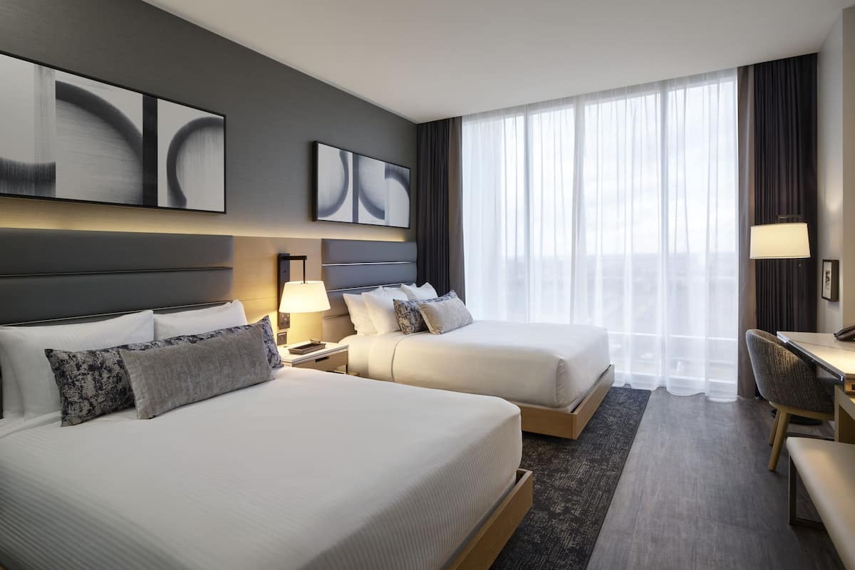 A hotel room with two beds, a desk, a chair, and a large window with curtains.