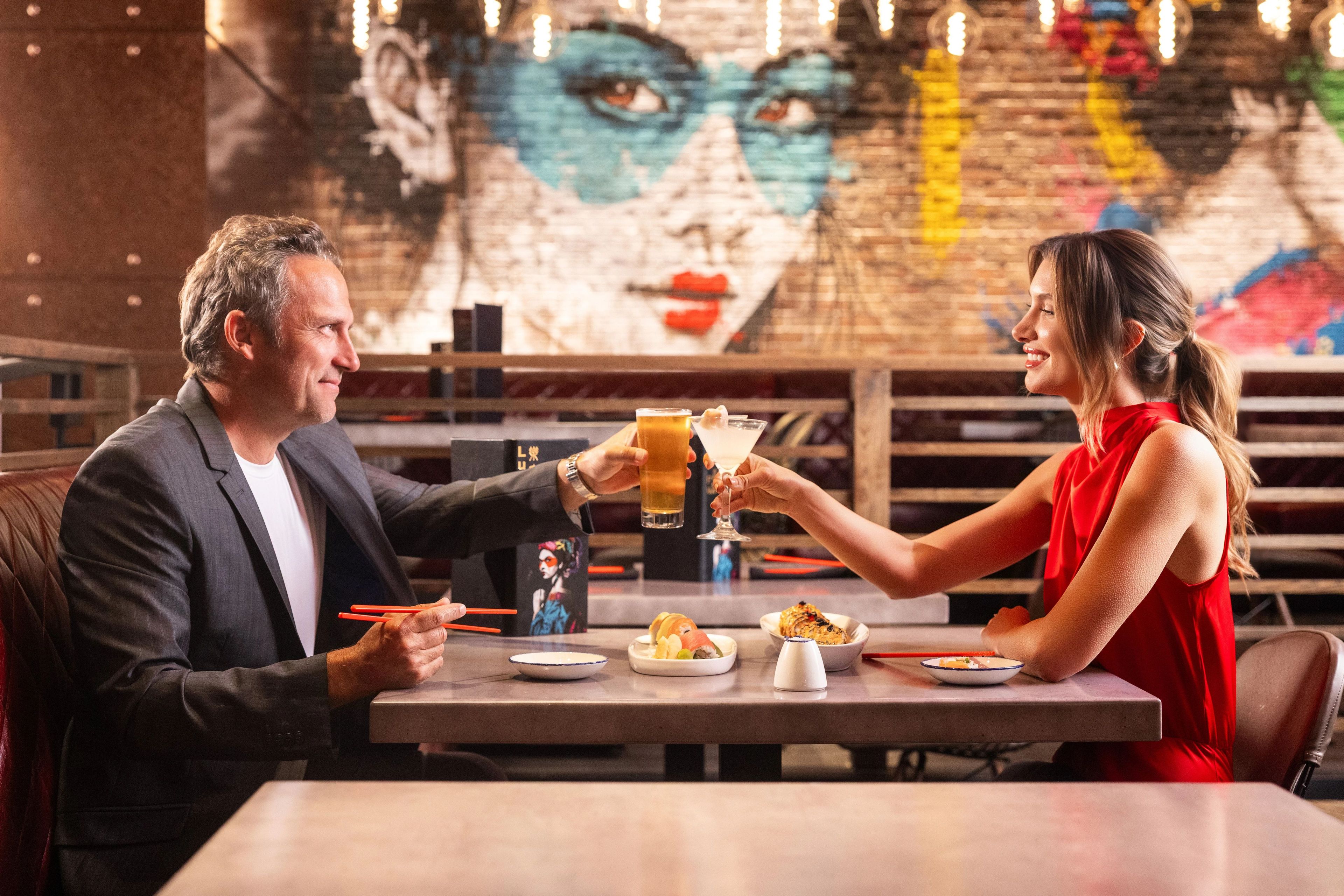 Un homme et une femme trinquent avec leurs boissons à une table dans un restaurant, une fresque murale servant de décor en arrière-plan.