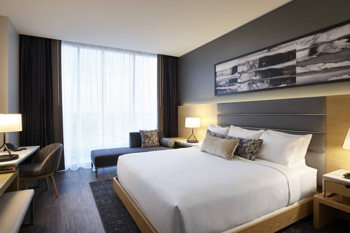 A modern hotel room with a large bed, a desk, a chair, and a view of the city through large windows.
