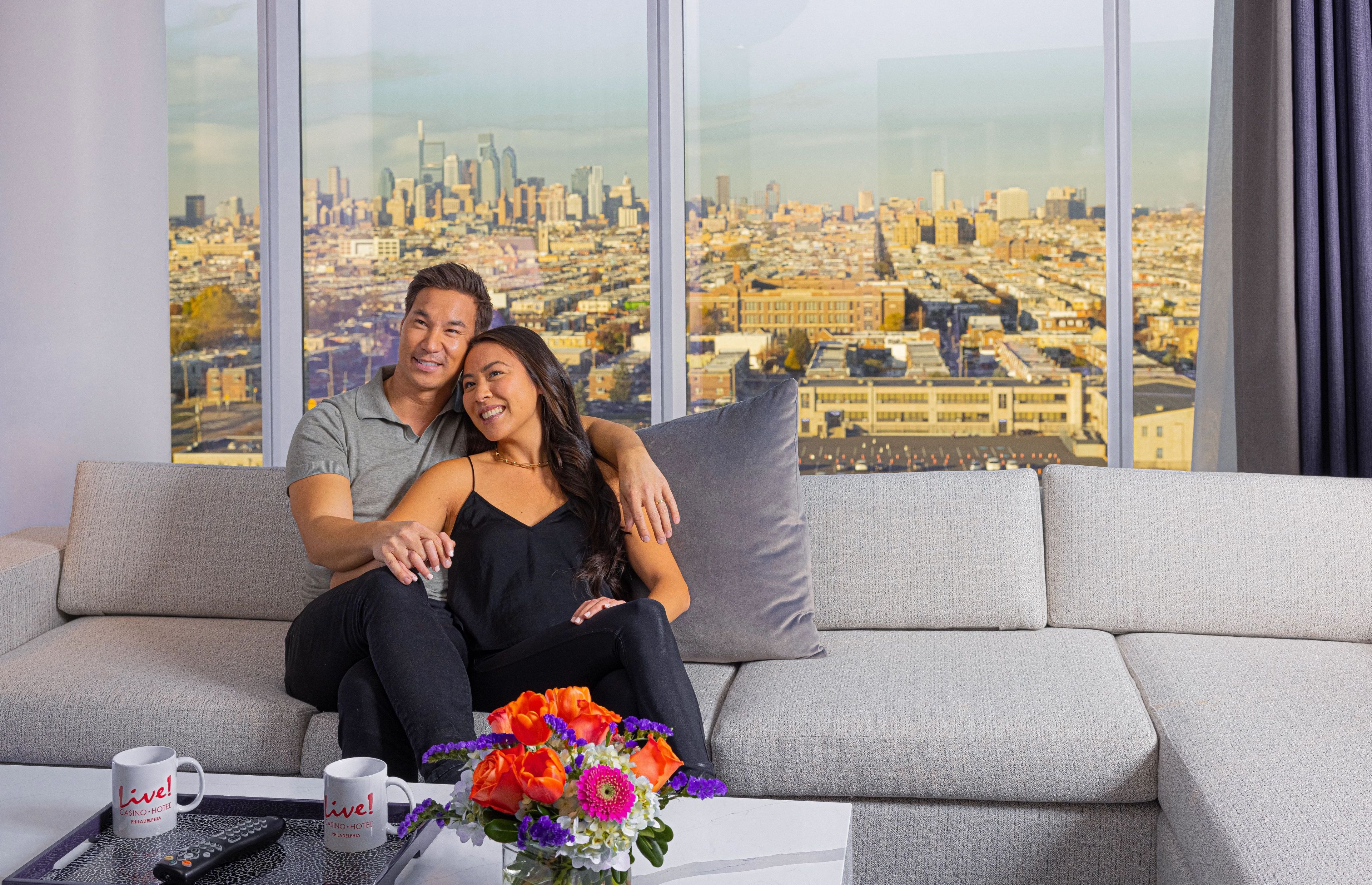 A couple sitting on a grey sofa with a cityscape view through large windows.