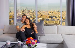 A couple sitting on a grey sofa with a cityscape view through large windows.