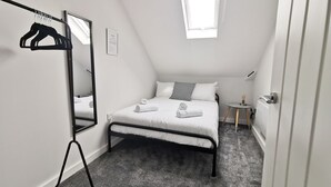 Une chambre à coucher comprenant un lit, un miroir, un portant à vêtements, une petite table avec une lampe et une fenêtre.