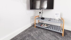 Un téléviseur fixé au mur, avec une étagère grise en dessous, sur laquelle reposent une télécommande et un magazine.