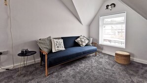 Une chambre de style loft moderne, avec un plafond en pente, un canapé bleu, des coussins à motifs, une petite table ronde et une grande fenêtre avec des stores.