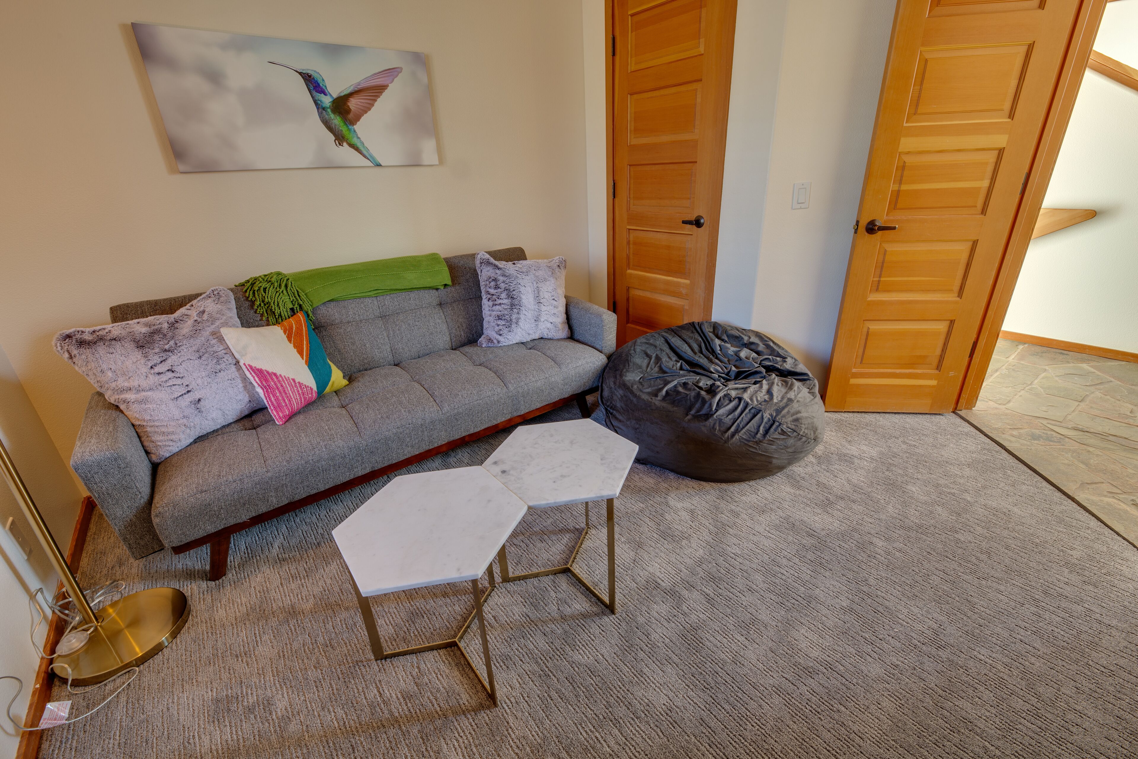 Un salón con un sofá gris, dos mesas blancas, un sillón informal y un cuadro de un colibrí.