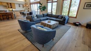 Amplia sala de estar con un sofá modular grande, dos sillones y una mesa de centro. Se ve una cocina al fondo, con armarios de madera y un comedor.