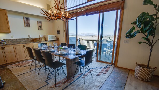Un comedor con una mesa dispuesta para cuatro personas, rodeada de sillas, una planta grande y vistas a través de puertas de cristal.