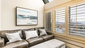 A modern living room with a leather sofa, a white ottoman, and a large window with blinds.