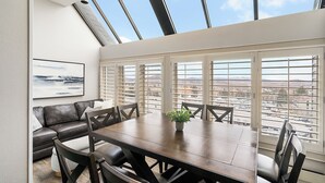 A dining area with a large table, chairs, a potted plant, and a view through large windows.